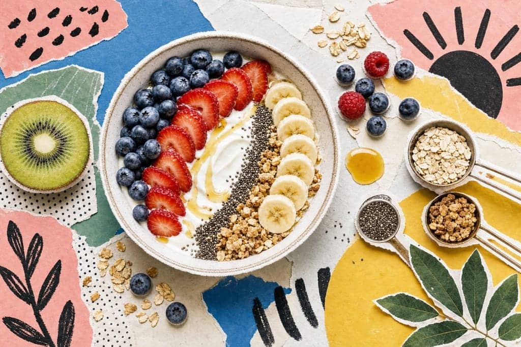 Editorial mixed-media collage of a ceramic breakfast bowl as the unmistakable central subject, filled with a meticulously organized calorie-conscious yogurt parfait shown from a crisp straight-down overhead angle, centered slightly left on a clean kitchen-counter inspired composition: thick white yogurt swirled smoothly into the bowl, topped in clearly separated arcs of fresh blueberries, sliced strawberries, banana coins, chia seeds, granola clusters, and a delicate drizzle of honey, with a few measuring-spoon ingredients scattered nearby for context. Surround the bowl with cut-out photographic elements of halved kiwi, loose oats, a tiny spill of berries, and subtle abstract nutrition-themed shapes, all layered over torn pastel paper and halftone scraps. Use a fresh, bright, motivating morning atmosphere with soft golden sunrise light, airy shadows, and a cheerful palette of creamy whites, berry reds, leaf greens, honey golds, and cool sky blue. Add bold flat colour shapes, hand-drawn marks, visible glue-stick texture, paper grain, and playful magazine composition. No characters, no humans, no text, no readable labels.
