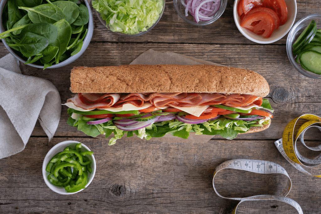 A photorealistic top-down view of a fresh, colorful Subway sandwich with vibrant layers of vegetables, cheese, and meats arranged neatly on a rustic wooden table, surrounded by small bowls of fresh ingredients and a clean measuring tape artfully coiled beside the sandwich, all under soft, natural lighting to evoke a mood of healthy lifestyle and mindful eating; no characters, text, or humans in the image.